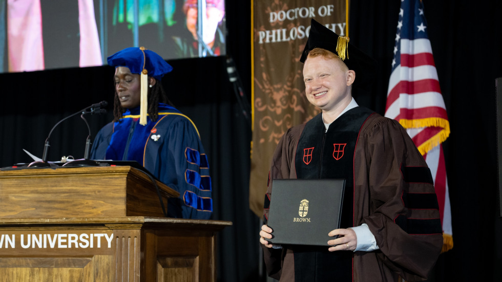 Gray Babbs, wearing Brown regalia, smiles holding an award certificate.