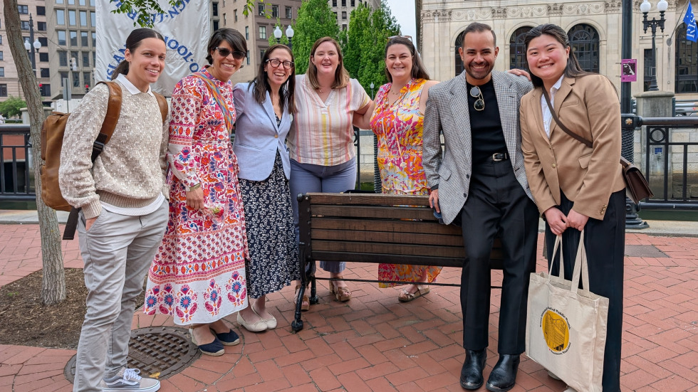 A group of faculty and students smiles outdoors with trees and urban riverwalk behind.